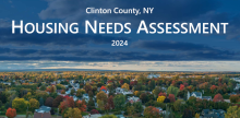 Birds eye view of houses in Clinton County with mountains in the background. A banner in white text reads Clinton County, NY Housing Needs Assessment 2024